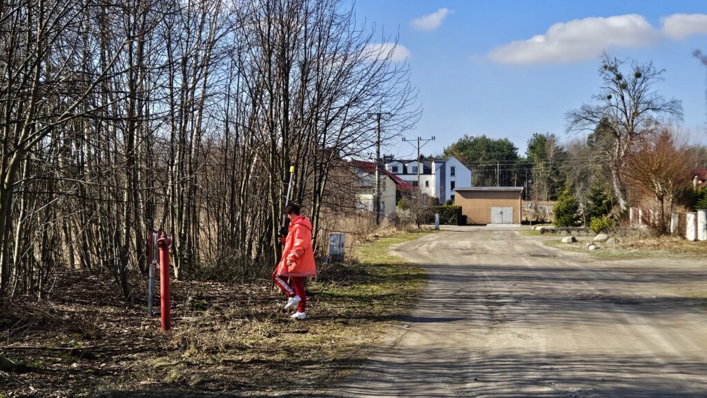Geodeta wykonujący pomiar GPS przy granicy działki w pobliżu drogi gruntowej i zabudowy jednorodzinnej – prace terenowe w zadrzewionym obszarze.
