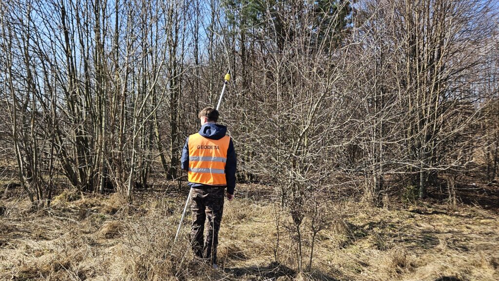 Geodeta wykonujący pomiar terenowy w zadrzewionym obszarze – praca z odbiornikiem GPS na działce porośniętej krzewami i drzewami.