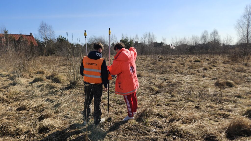 Geodeta wykonujący pomiary terenowe na działce – pomiar GPS przy użyciu tyczek geodezyjnych na niezabudowanym terenie pod inwestycję budowlaną.
