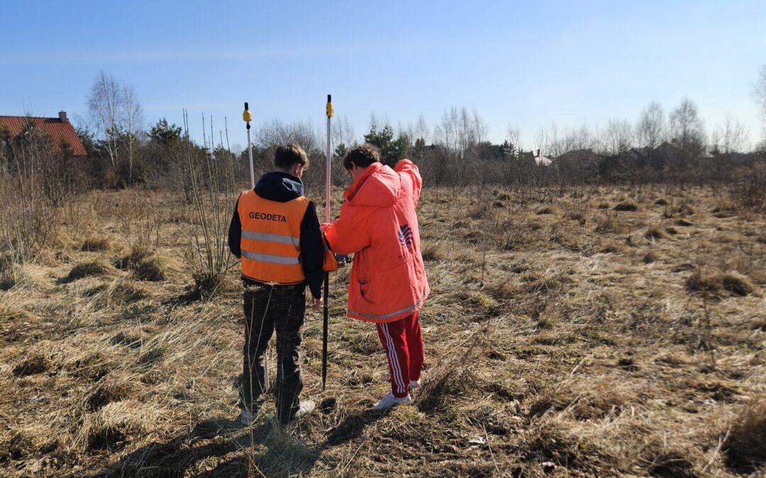 Geodeta wykonujący pomiary terenowe na działce – pomiar GPS przy użyciu tyczek geodezyjnych na niezabudowanym terenie pod inwestycję budowlaną.