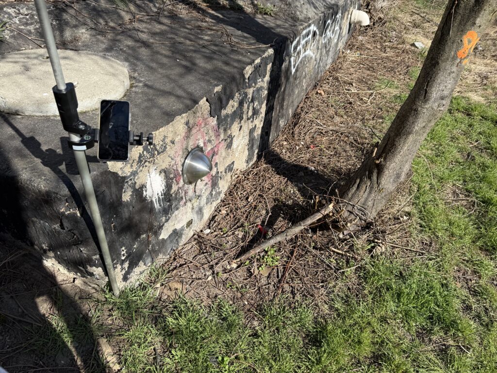 Geodezyjny pomiar terenowy przy murze – tyczka pomiarowa ze smartfonem, punkt odniesienia na ścianie oraz oznaczenia na drzewie wskazujące zakres prac.